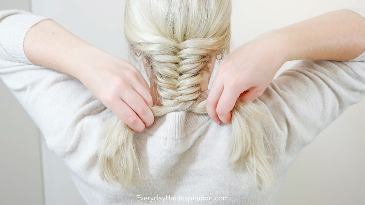 Blonde woman braiding her hair into a fishtail braid down the back of the head. 