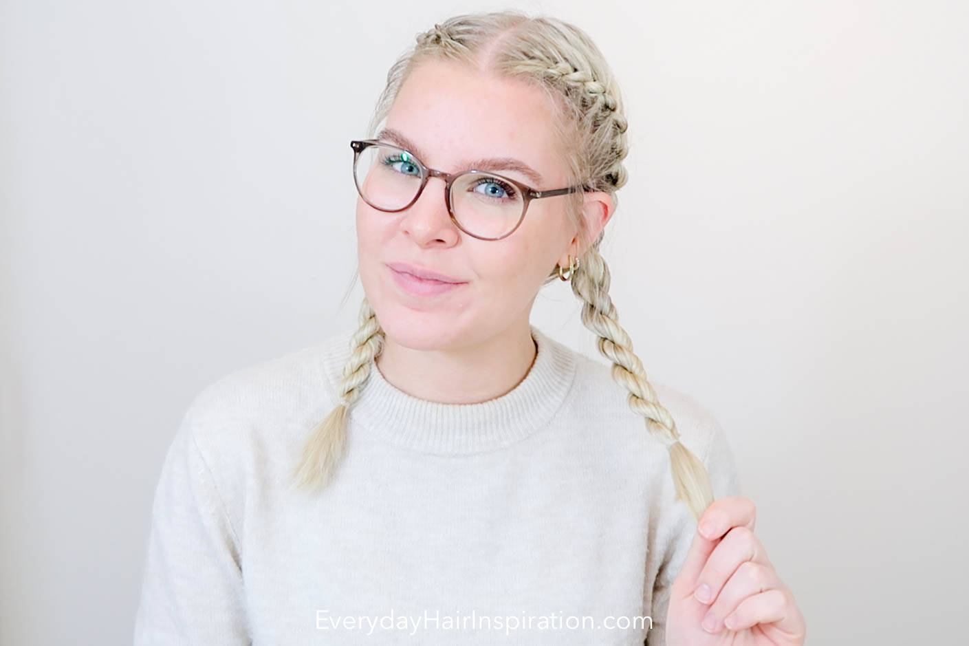 Blonde girl seen from the front, looking into the camera with her hair braided into double French rope braids, holding one braid in her hand. 